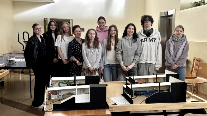 Studenti ar pasniedzēju pozē aiz galda ar pabeigtiem interjera un konstrukciju modeļiem.