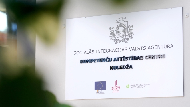 Balta plāksne pie sienas ar Latvijas valsts ģerboni un uzrakstu "SOCIĀLĀS INTEGRĀCIJAS VALSTS AĢENTŪRA, KOMPETENČU ATTĪSTĪBAS CENTRS, KOLEDŽA", ko papildina Eiropas Savienības, Nacionālā attīstības plāna 2027 un SIVA logotipi.