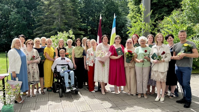 SIVA koledžas absolventi ar pasniedzējiem svinīgā izlaidumā ārpus telpām. Cilvēki smaida, turot ziedus un tumši zaļus diplomus.