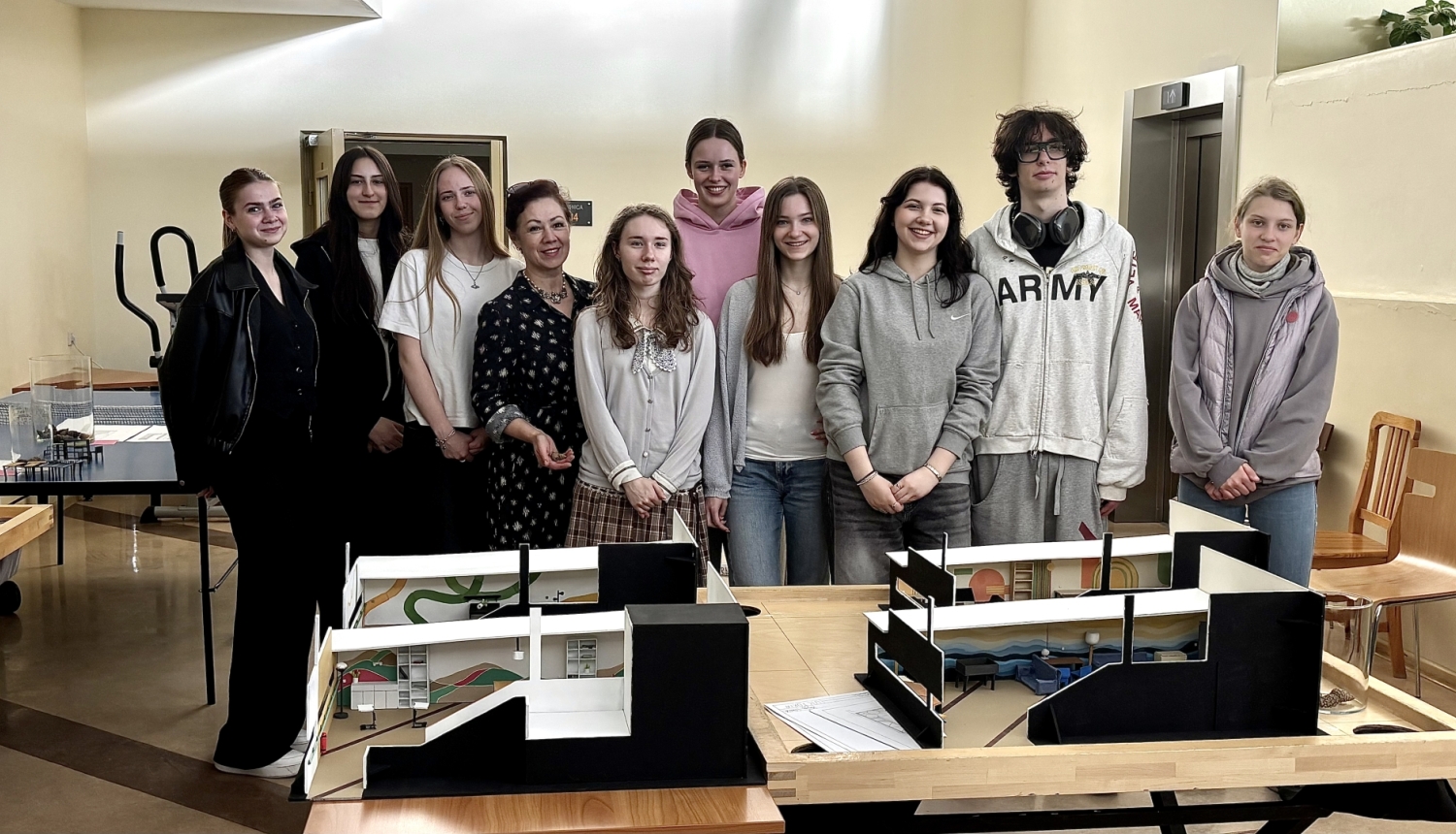 Studenti ar pasniedzēju pozē aiz galda ar pabeigtiem interjera un konstrukciju modeļiem.