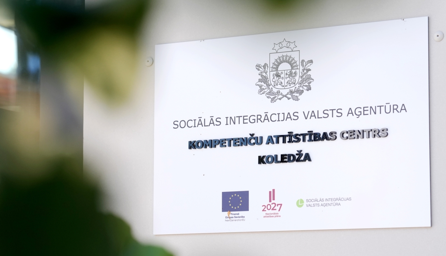 Balta plāksne pie sienas ar Latvijas valsts ģerboni un uzrakstu "SOCIĀLĀS INTEGRĀCIJAS VALSTS AĢENTŪRA, KOMPETENČU ATTĪSTĪBAS CENTRS, KOLEDŽA", ko papildina Eiropas Savienības, Nacionālā attīstības plāna 2027 un SIVA logotipi.
