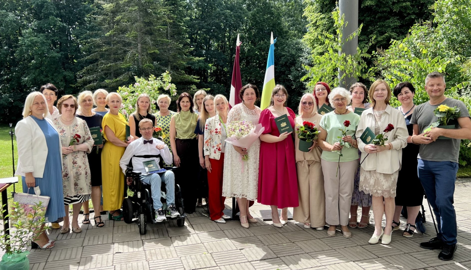 SIVA koledžas absolventi ar pasniedzējiem svinīgā izlaidumā ārpus telpām. Cilvēki smaida, turot ziedus un tumši zaļus diplomus.