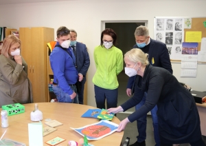 SIVA darbinieces un viesi atrodas sociālā rehabilitētāja nodarību telpā. Uz galda novietoti dažādi sociālālās rehabilitācijas nodarbībās tapuši darbiņi. Sociālais rehabilitētājs uz galda novieto vēl divus aplikāciju darbus.
