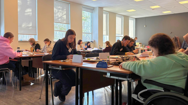 2.	Cilvēki sēž pie galda un strādā ar rokdarbiem. Telpā ir lieli logi un daudz gaismas.