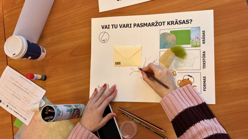 3.	Cilvēks pie galda zīmē uz lapas. Uz galda ir krāsas, papīrs un citi radoši materiāli.