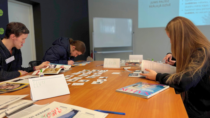 4.	Trīs cilvēki sēž pie galda un strādā ar papīru un attēliem. Uz galda ir kartītes, grāmatas un līme.