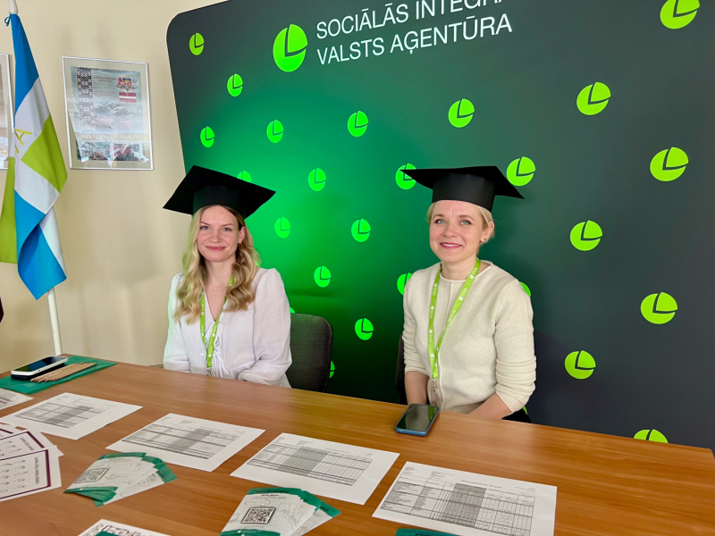 SIVA pārstāves akadēmiskajās cepurēs pie informatīvā stenda.