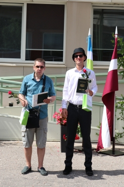 Divi absolventi - puiši - stāv kājās, rokās turot iegūto diplomu, ziedus un dāvanas