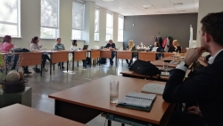 Plašā telpā sēž cilvēki pie galdiem