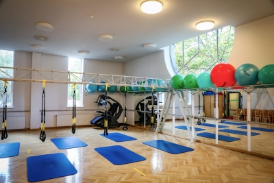 Fizioterapijas lielā zāle, kurā uz grīdas ir zili vingrošanas paklāji, uz sienām spoguļi, uz plauktiem stāv dažāda izmēra un lieluma vingrošanas bumbas utt.