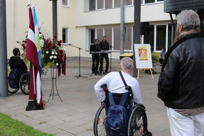 Ansambļa vadītājs un viens audzēknis kopā spēlē sintezatoru. Redzami tuvumā esošie pasākuma dalībnieki, Latvijas valsts karogs un SIVA karogs, kā arī ziedu kompozīcija