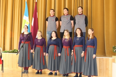 Jaunieši tautastērpos stāv pie skatuves, fonā redzams gan SIVA izglītības iestāžu karogs, gan Latvijas valsts karogs