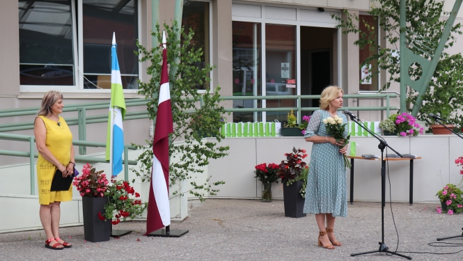 SIVA direktore I.Jurševska tur rokās baltas rozes un saka klātesošajiem runu. Fonā redzams Latvijas karogs un SIVA karogs, kā arī ziedi un absolventiem paredzētās dāvanas