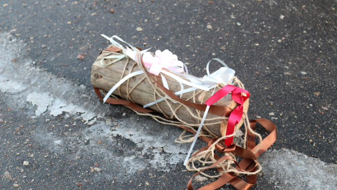 Uz asfalta novietots koka bluķis, kas izgreznots ar baltām, brūnām, sarkanām un rozā lentītēm.