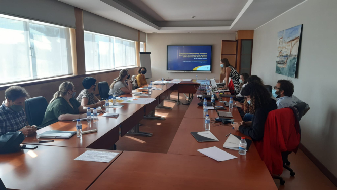 Konferenču telpā ap galdiem sēž cilvēki un skatās uz ekrāna esošo prezentāciju