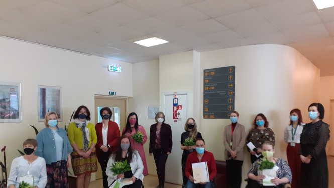 Jūrmalas profesionālās vidusskolas absolventi, kas ieguvuši Šuvēja palīga kvalifikāciju, sēž krēslos, turot rokā maijpuķītes, izglītības dokumentus un dāvanas. Aiz viņiem kājās stāv skolas personāls. Visiem klātesošajiem ir uzvilktas sejas maskas.