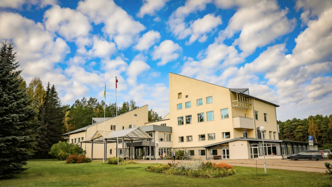 Jūrmalas profesionālās vidusskolas dzeltenīgā ēka, apkārt skujkoki, zilas debesis ar baltiem mākonīšiem