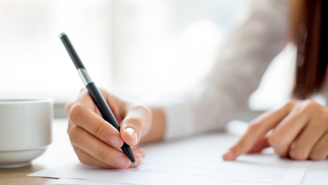 Ilustratīvs attēls - uz galda stāv baltas papīra lapas. Uz vienas no tām tiek rakstīts ar pildspalvu.