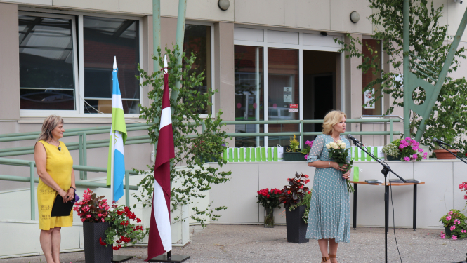 SIVA direktore I.Jurševska tur rokās baltas rozes un saka klātesošajiem runu. Fonā redzams Latvijas karogs un SIVA karogs, kā arī ziedi un absolventiem paredzētās dāvanas