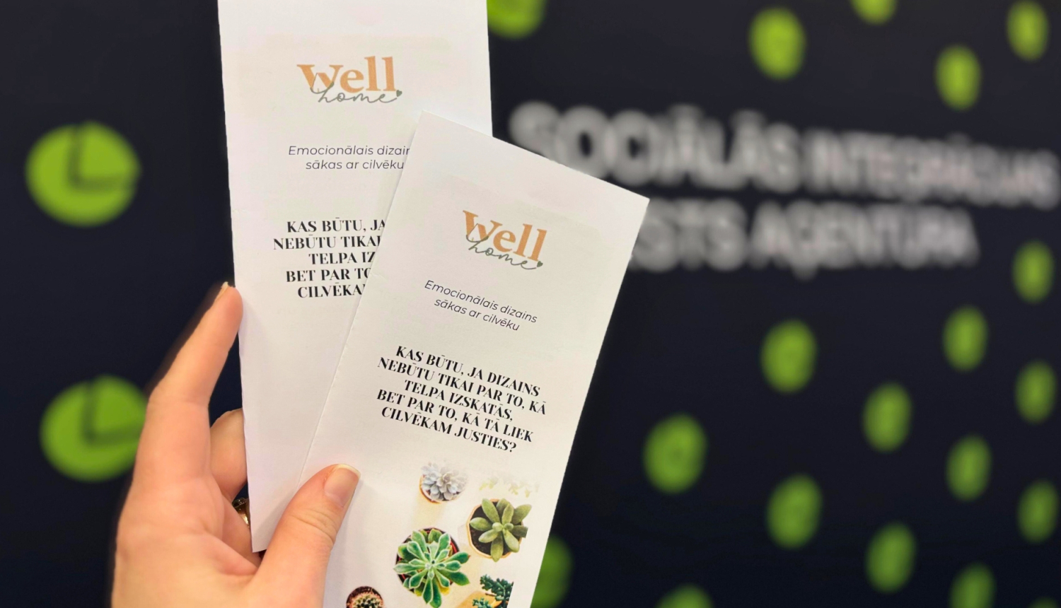 1.	Roka tur divus bukletus ar uzrakstu “Well home”. Uz bukletiem ir teksts par emocionālo dizainu un mazi augi attēla apakšā.