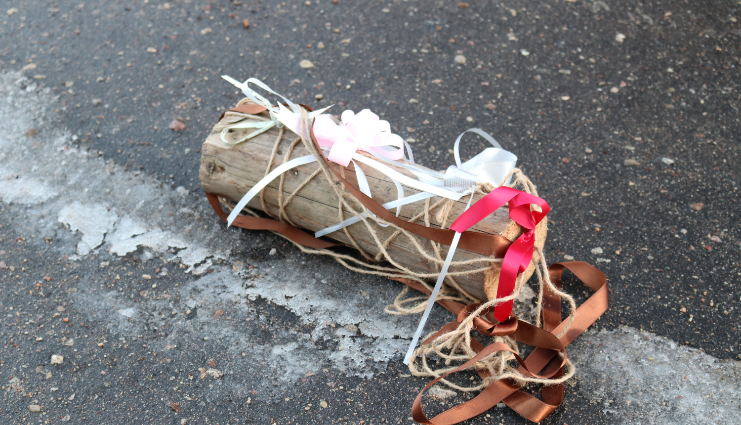 Uz asfalta novietots koka bluķis, kas izgreznots ar baltām, brūnām, sarkanām un rozā lentītēm.