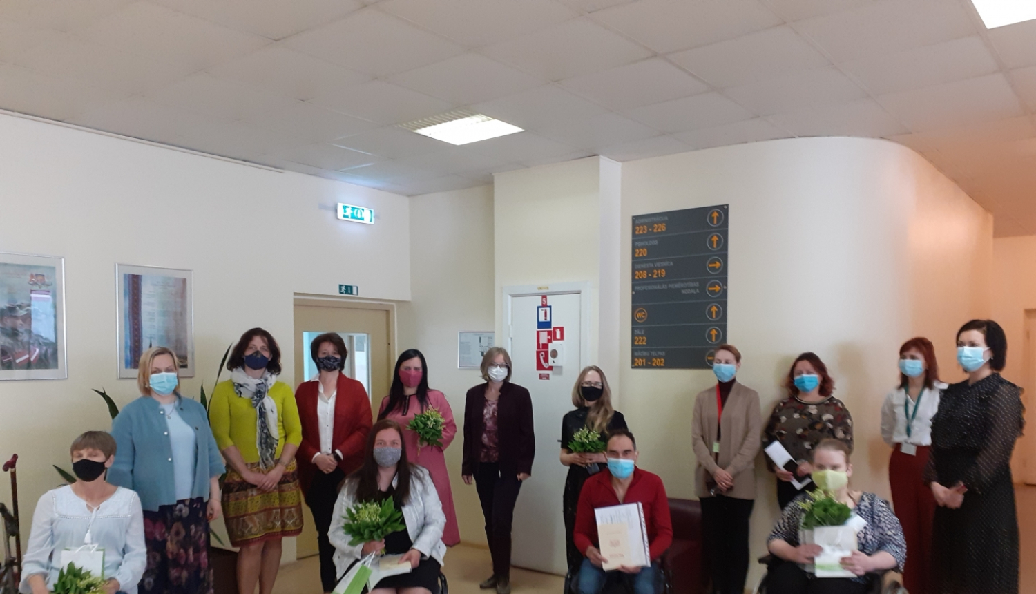 Jūrmalas profesionālās vidusskolas absolventi, kas ieguvuši Šuvēja palīga kvalifikāciju, sēž krēslos, turot rokā maijpuķītes, izglītības dokumentus un dāvanas. Aiz viņiem kājās stāv skolas personāls. Visiem klātesošajiem ir uzvilktas sejas maskas.