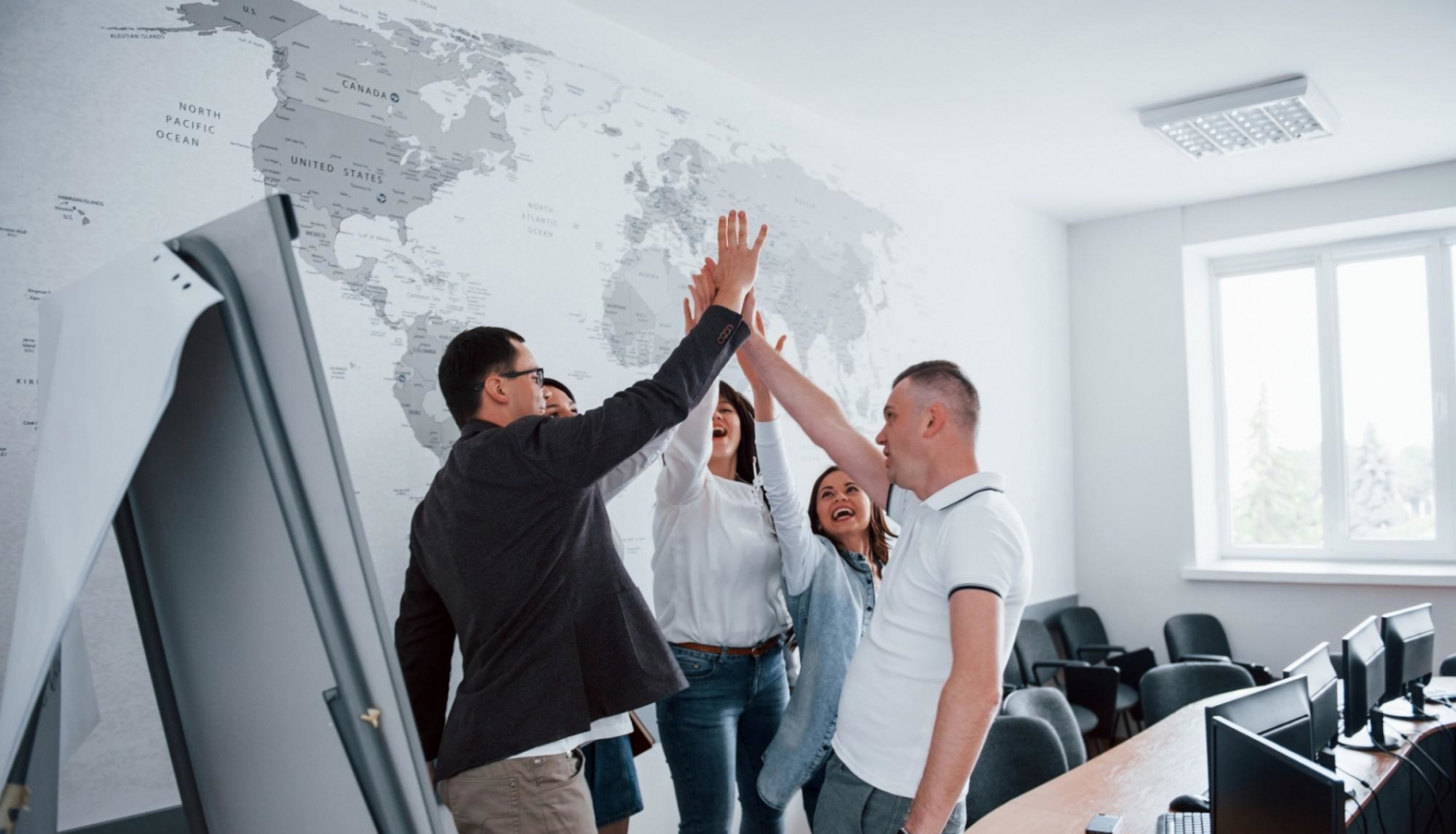 Pieci priecīgi jaunieši un jaunietes stāv telpas vidū un, katrs pacēlis vienu roku gaisā, visi kopā "dod pieci!"