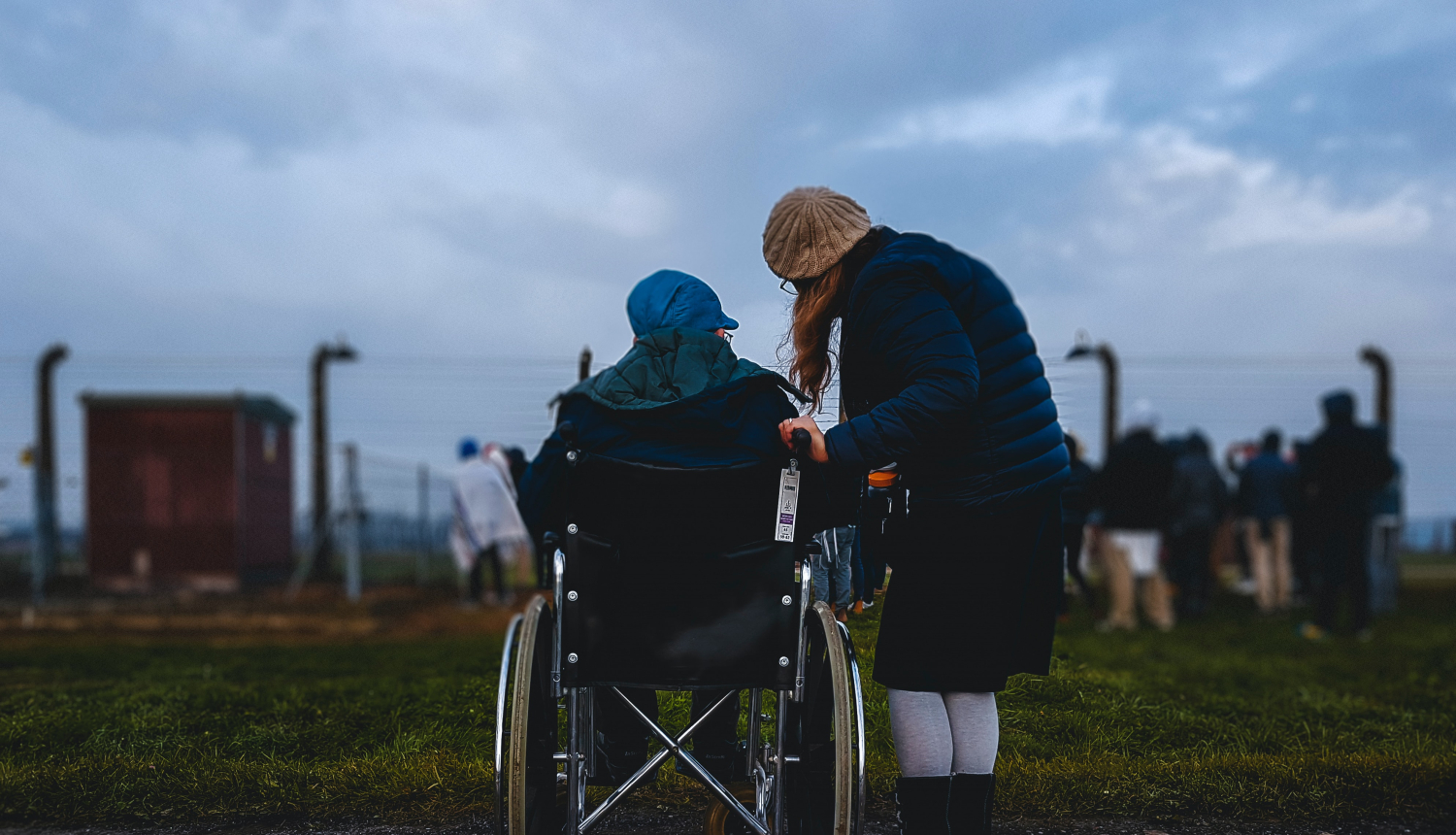 Sieviete stāv nedaudz pieliekusies pie vīrieša, kas atrodas ratiņkrēslā