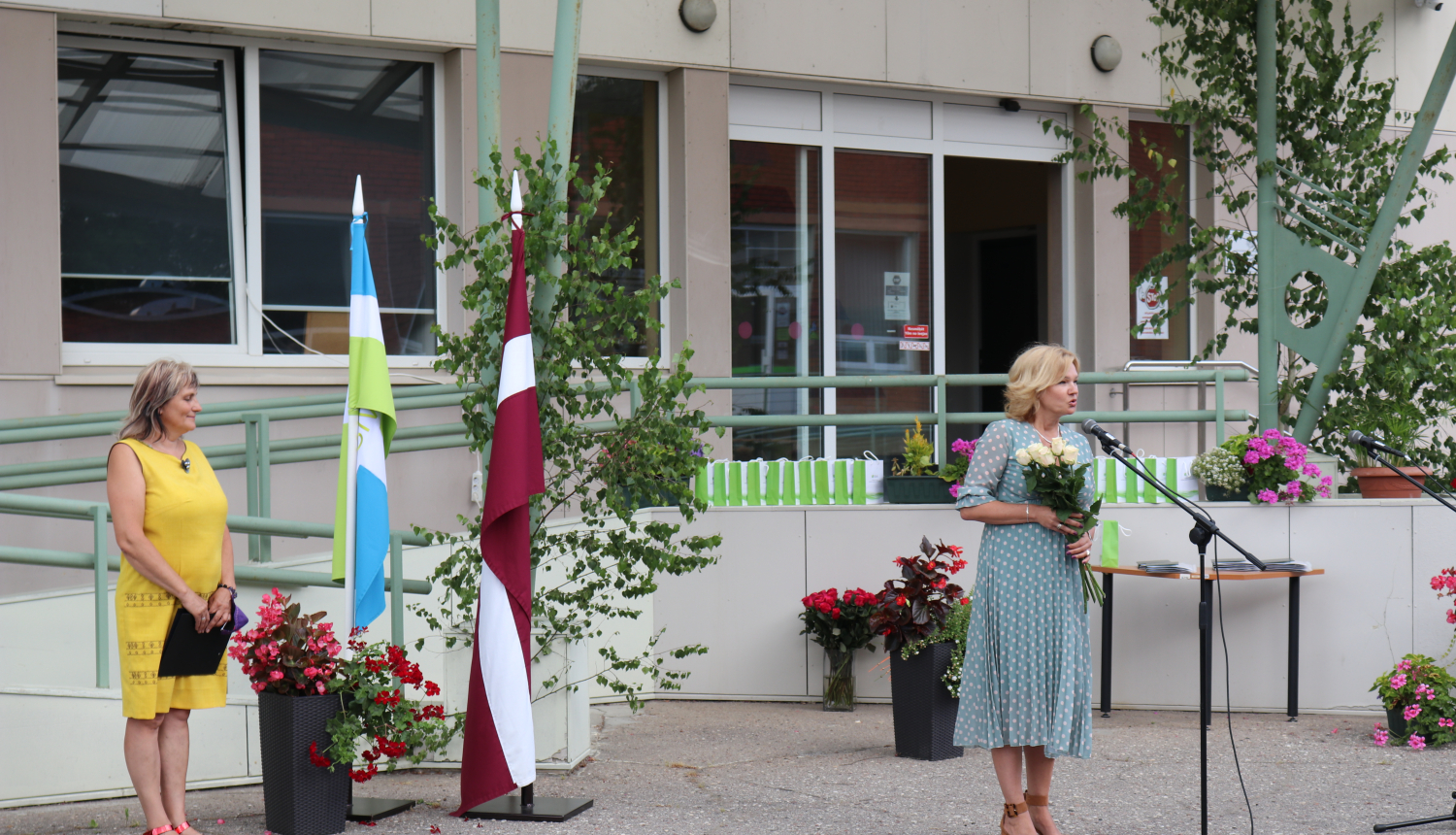 SIVA direktore I.Jurševska tur rokās baltas rozes un saka klātesošajiem runu. Fonā redzams Latvijas karogs un SIVA karogs, kā arī ziedi un absolventiem paredzētās dāvanas