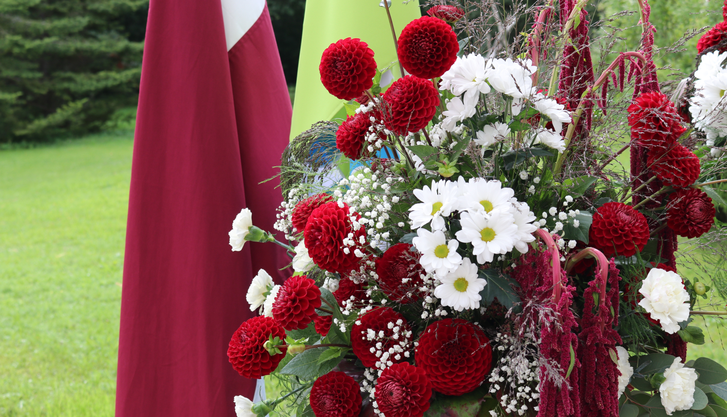 Sarkanu un baltu ziedu kompozīcija. Fonā redzams Latvijas Republikas karogs un SIVA karogs